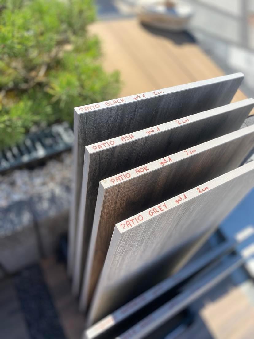 Przekrój czterech próbek płytek tarasowych Patio w odcieniach szarości, czerni i brązu, każda z czerwoną etykietą z nazwą i specyfikacją, ustawionych pod kątem na zewnątrz w słoneczny dzień.