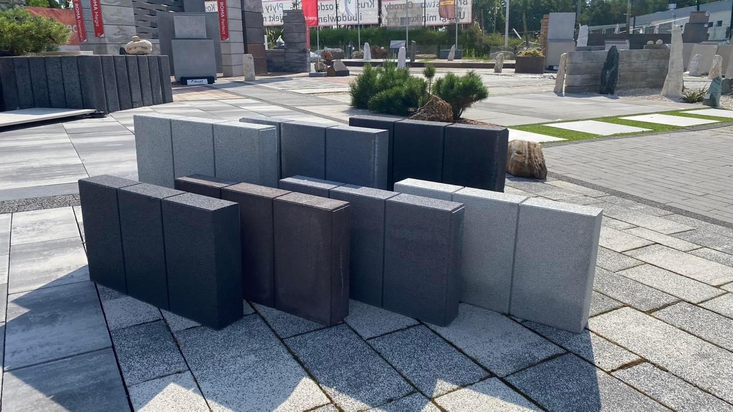 Ekspozycja różnokolorowych, prostokątnych bloków betonowych na placu z kostki brukowej, prezentacja różnych odcieni szarości i czerni, z elementami zieleni w tle.