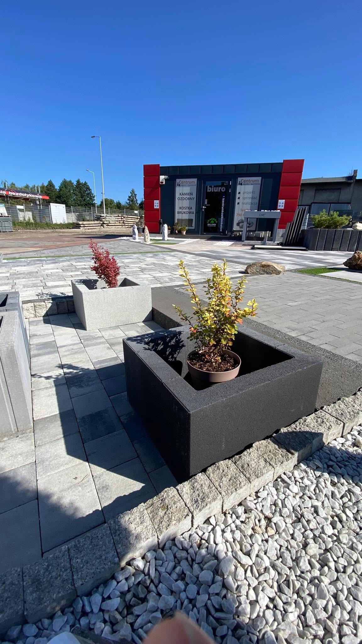 Ekspozycja kostki brukowej i granitowej przed biurem firmy, z donicami z roślinami, prezentująca różne wzory i kolory nawierzchni.