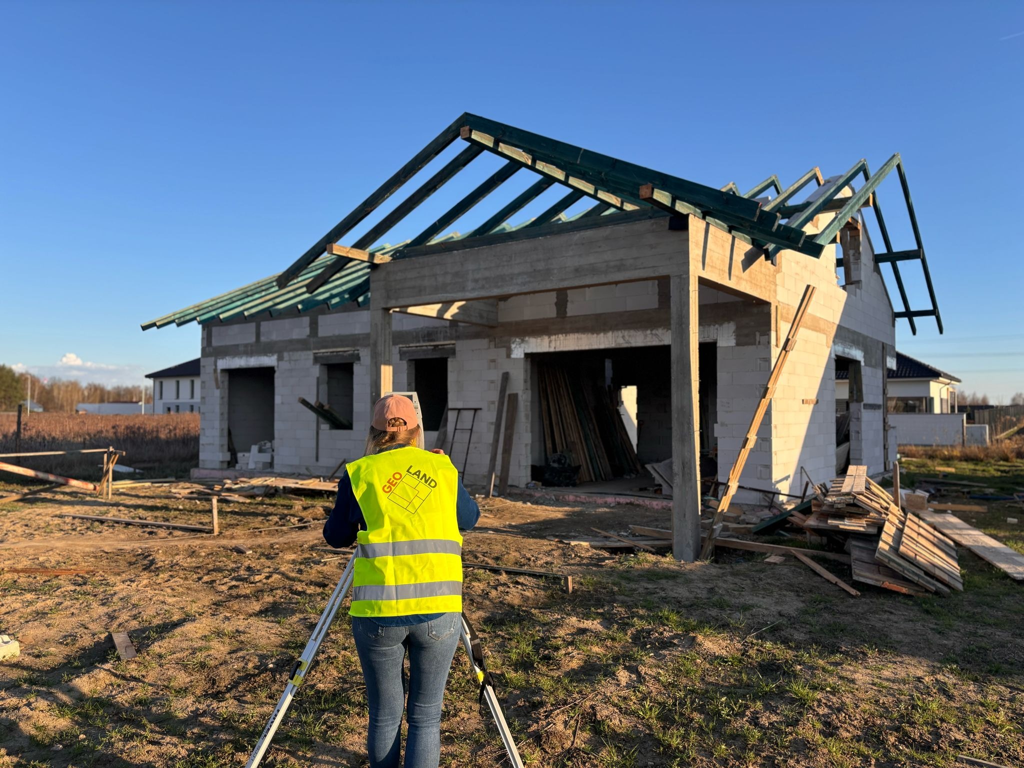 Geodeta w żółtej kamizelce 'GEO LAND' mierzy teren pod budowę domu jednorodzinnego w stanie surowym otwartym, używając niwelatora.