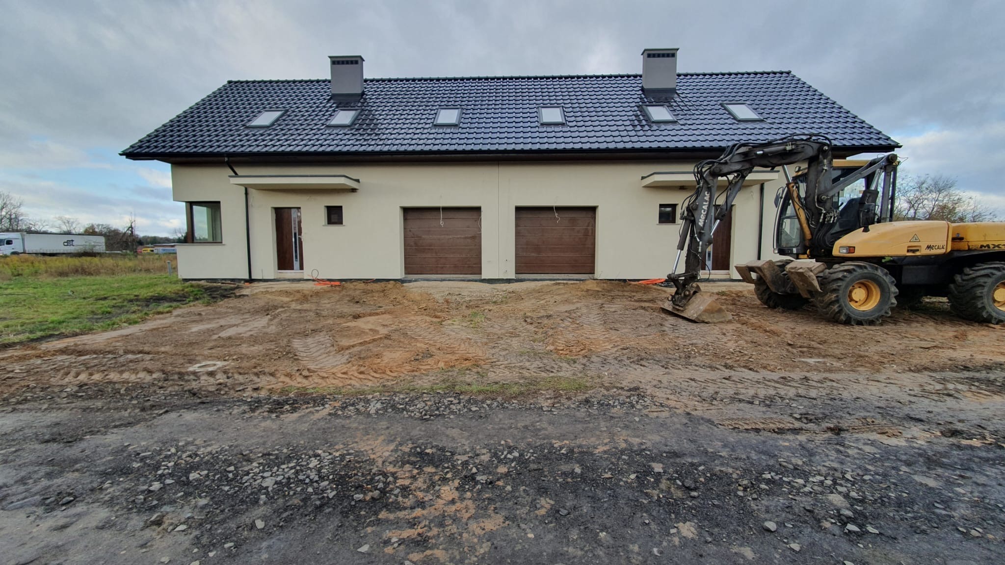 Dwukondygnacyjny dom z ciemną dachówką i beżową elewacją w trakcie prac ziemnych z użyciem koparki Mecalac MX1, widok z przodu na plac budowy.