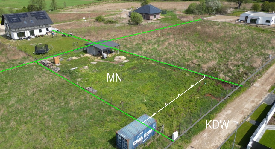 Zdjęcie z drona przedstawia wytyczony teren działki z oznaczeniami MN i KDW, otoczonej budynkami mieszkalnymi i polami. Widoczny kontener i elementy infrastruktury.