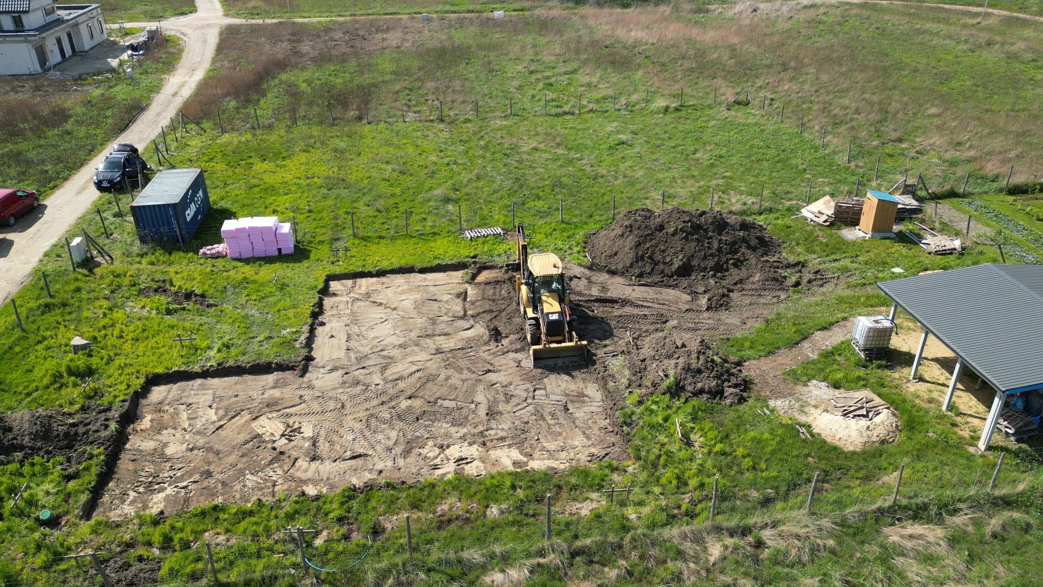 Żółta koparka CAT wyrównuje teren pod budowę, widok z góry. Na działce kontener, izolacja i wykopane fundamenty. W tle budynek i droga.