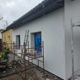 DAWBUD - Świeżo wyszpachlowana ściana domu z rusztowaniem, drabina oparta o ścianę, okna zabezpieczone folią. Widoczne narzędzia i materiały budowlane.