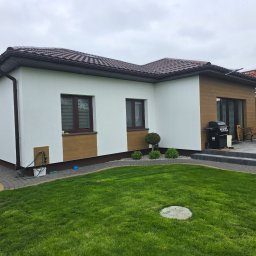 DAWBUD - Biała elewacja domu jednorodzinnego z brązowymi akcentami, widok z ogrodu. Uwagę zwraca kontrast kolorów i zadbany trawnik. Widoczny grill i meble ogrodowe.