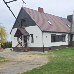 DAWBUD - Biały dom z brązowym dachem i elementami elewacji, widok z zewnątrz, trawnik i podjazd. W tle drzewa i niebo. Wieniec na drzwiach wejściowych.