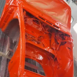 Pomarańczowa karoseria samochodu w trakcie lakierowania, widoczna na metalowej kratownicy w lakierni, z logo 'irx karosserie' w tle.