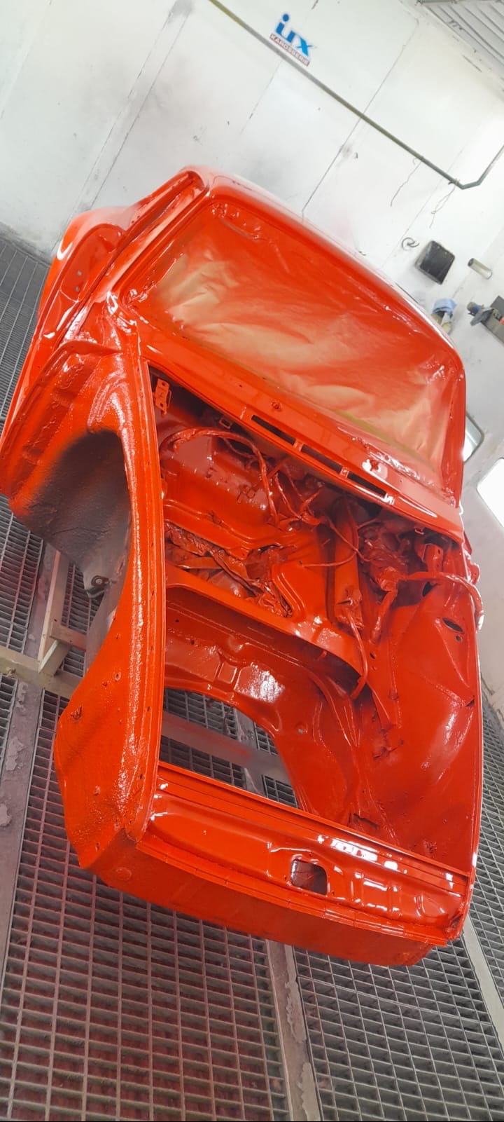 Pomarańczowa karoseria samochodu w trakcie lakierowania, widoczna na metalowej kratownicy w lakierni, z logo 'irx karosserie' w tle.