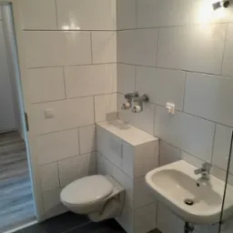 Nowoczesna łazienka z białymi, prostokątnymi płytkami ściennymi, wiszącą toaletą z zabudowaną spłuczką, umywalką nablatową i ciemnoszarą podłogą z płytek.