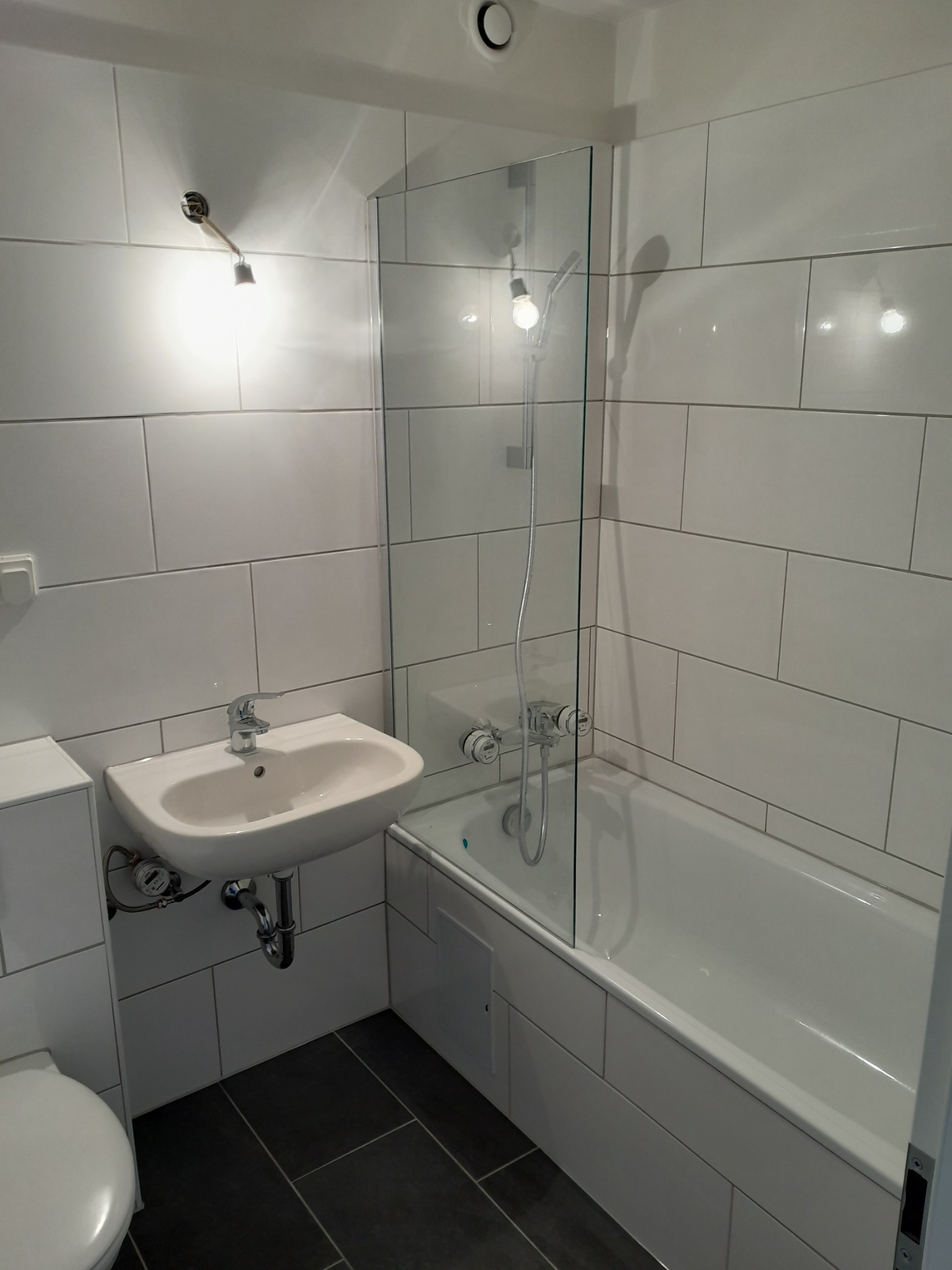 Nowoczesna łazienka z białymi, prostokątnymi płytkami, czarną podłogą, białą umywalką nablatową i wanną z przeszklonym parawanem.