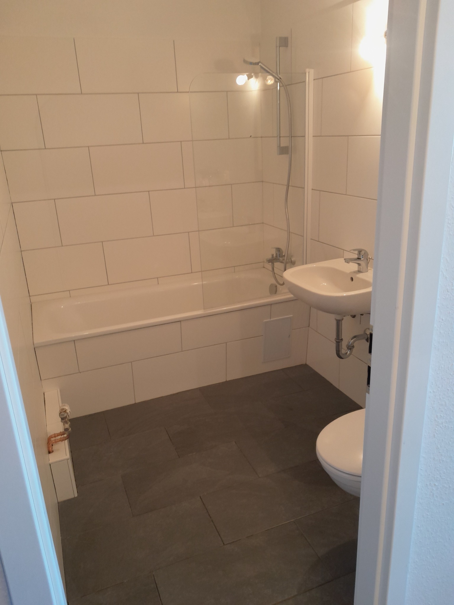 Nowoczesna łazienka z białymi, prostokątnymi płytkami, szarą podłogą, wanną z parawanem, umywalką i toaletą, widziana zza framugi drzwi.