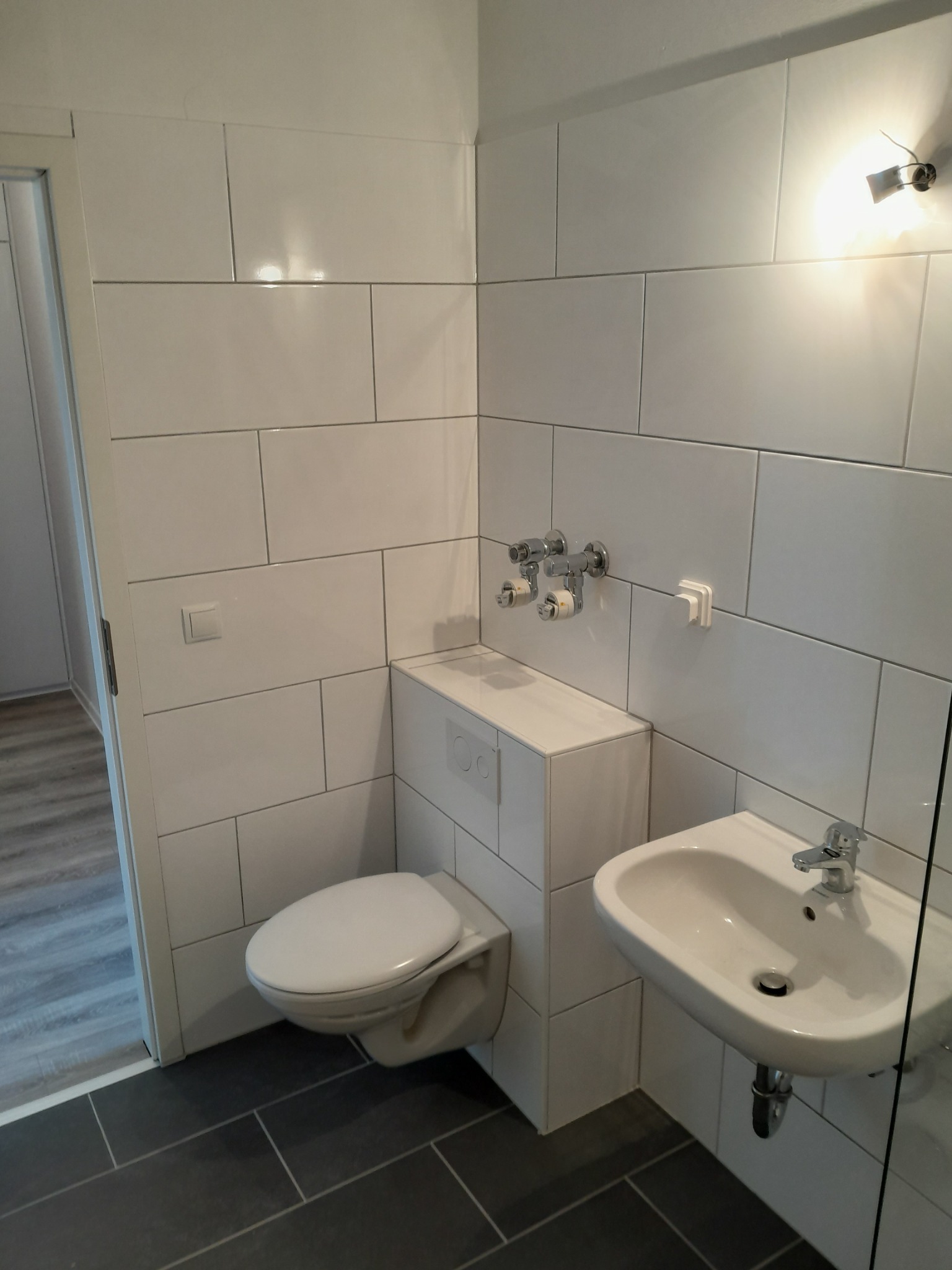 Nowoczesna łazienka z białymi, prostokątnymi płytkami ściennymi, wiszącą toaletą z zabudowaną spłuczką, umywalką nablatową i ciemnoszarą podłogą z płytek.