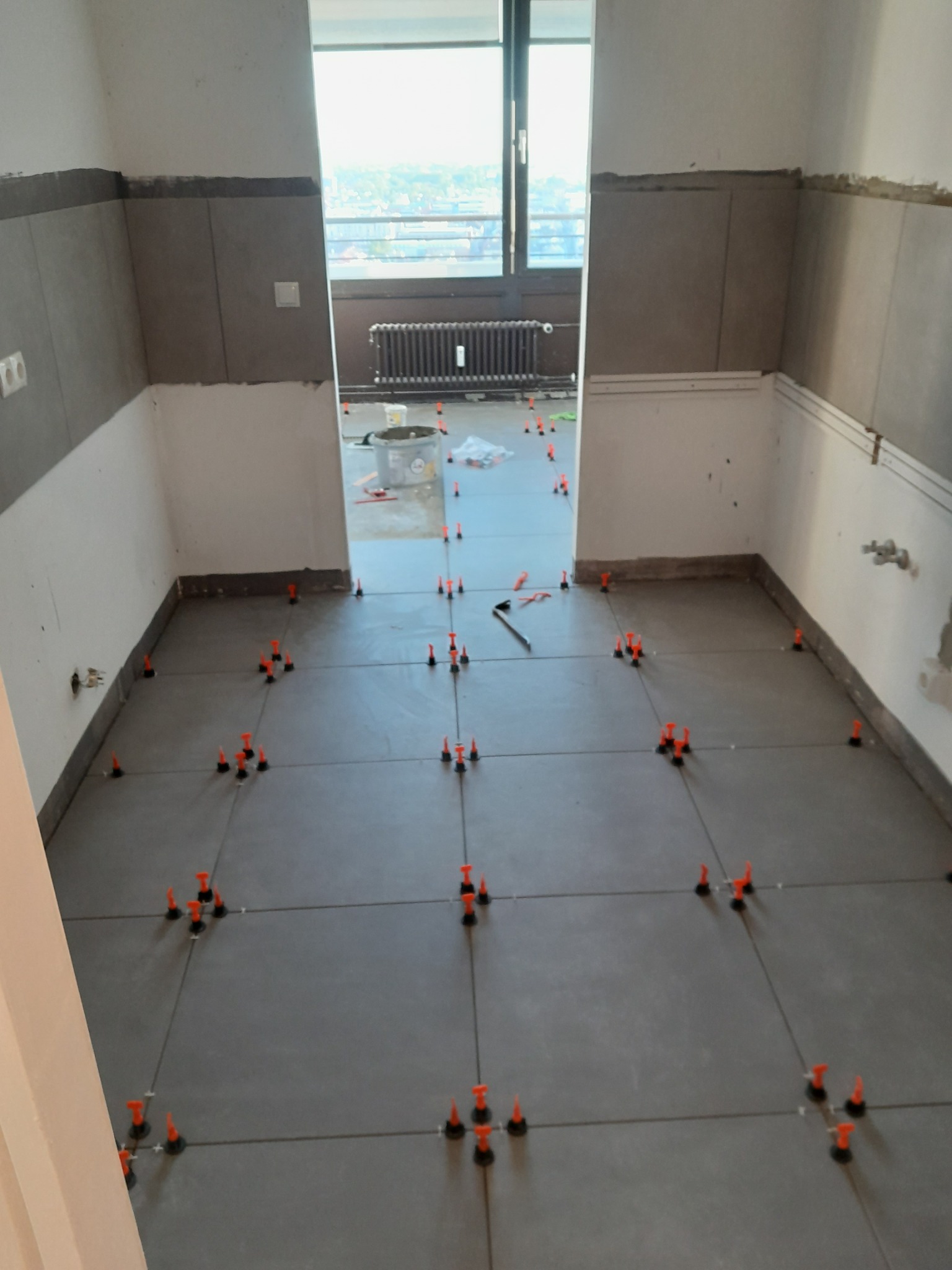 Szare płytki podłogowe w trakcie układania z użyciem pomarańczowych klipsów poziomujących, widoczne narzędzie do cięcia płytek i wiadro z zaprawą w tle.