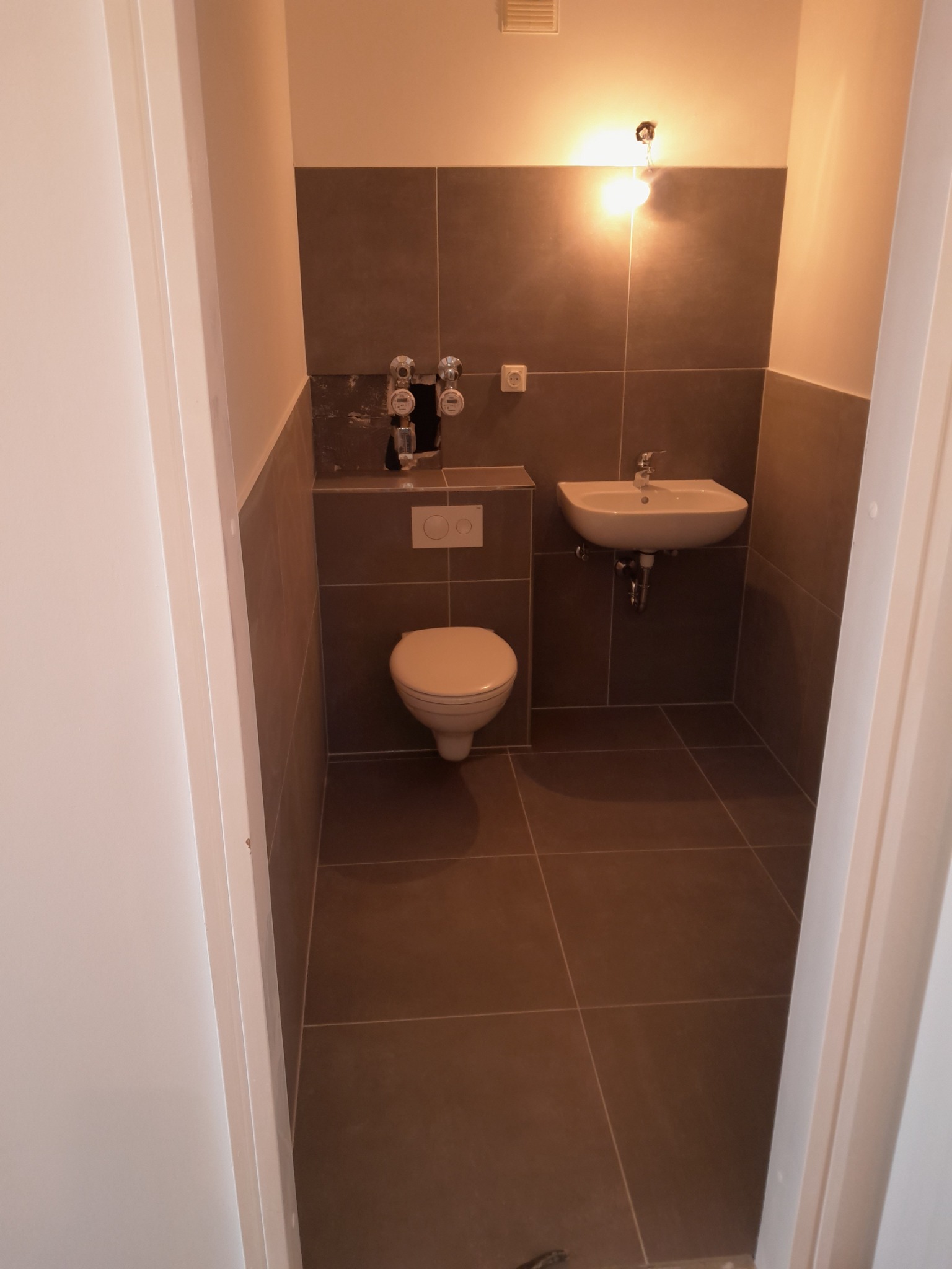 Nowoczesna łazienka z brązowymi płytkami, wiszącą umywalką i toaletą, widok zza uchylonych białych drzwi.