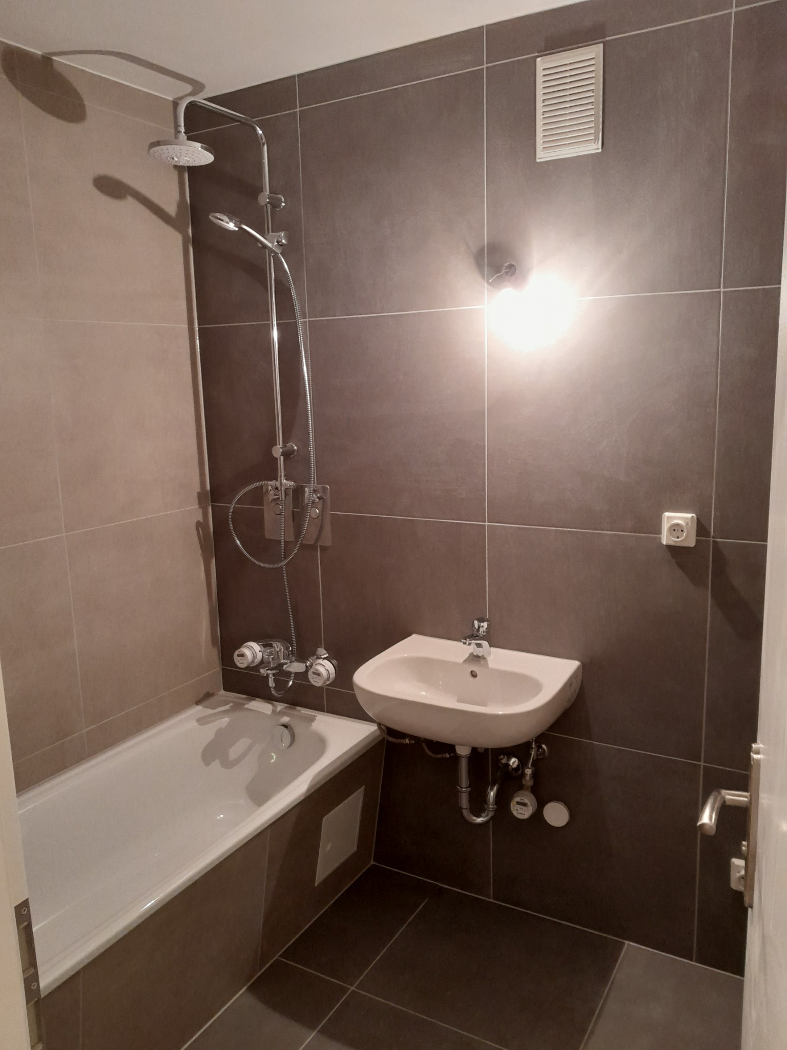 Nowoczesna łazienka z brązowymi płytkami, białą wanną i umywalką wiszącą, widoczne rury odpływowe, chromowana armatura i wentylator w ścianie.