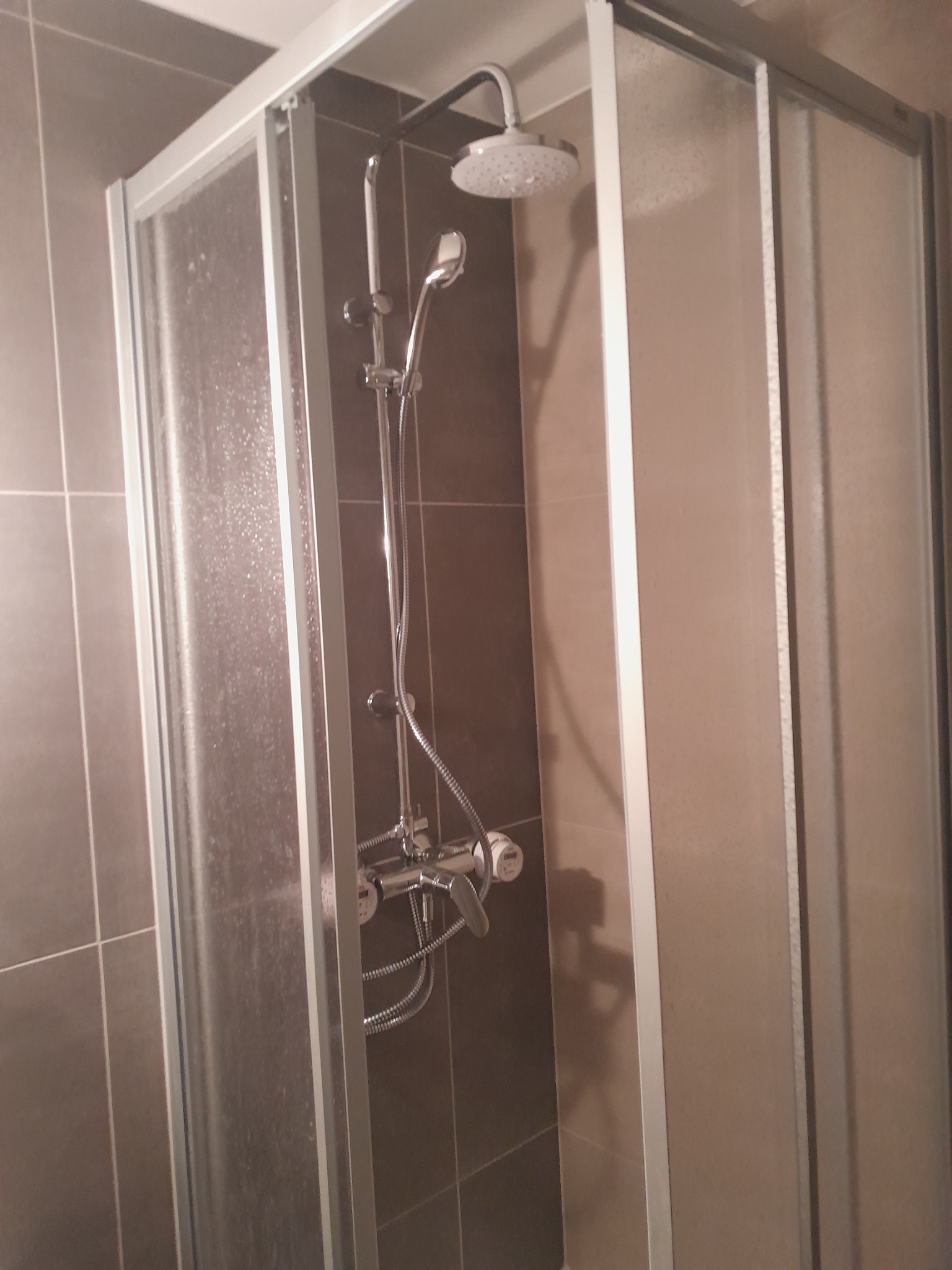 Szklana kabina prysznicowa z chromowaną armaturą, zamontowana na brązowych płytkach ściennych. Na szybie widoczne ślady po wodzie.