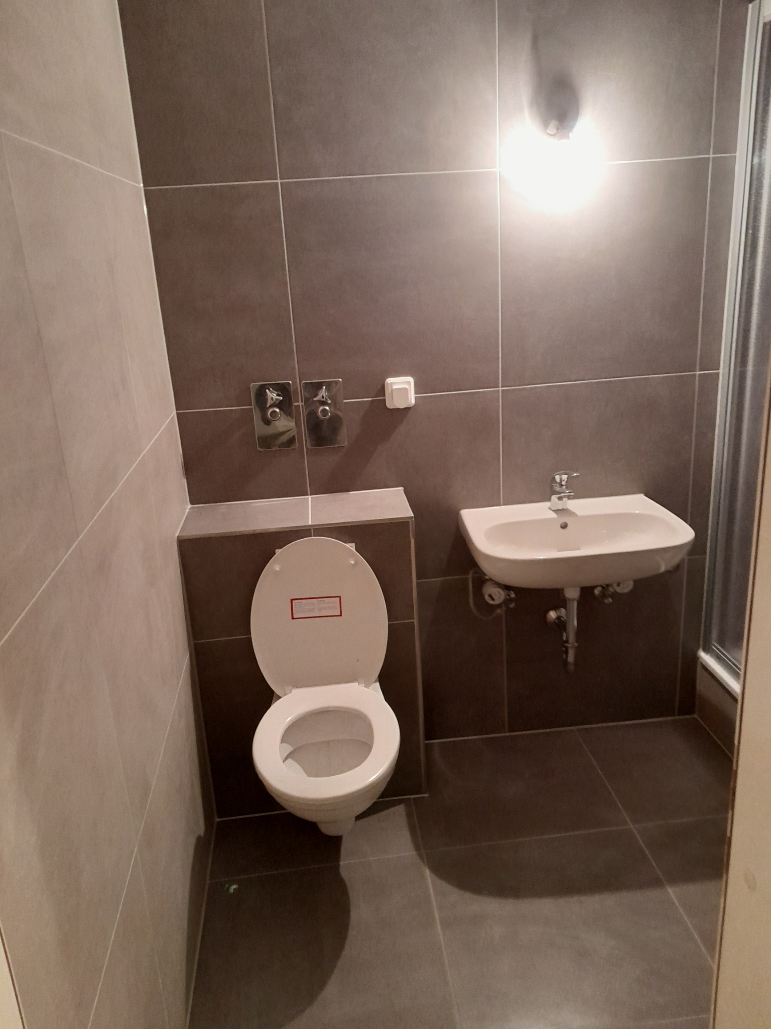 Nowoczesna łazienka z szarymi płytkami, wiszącą białą umywalką i toaletą z otwartą deską, widoczne chromowane baterie i oświetlenie sufitowe