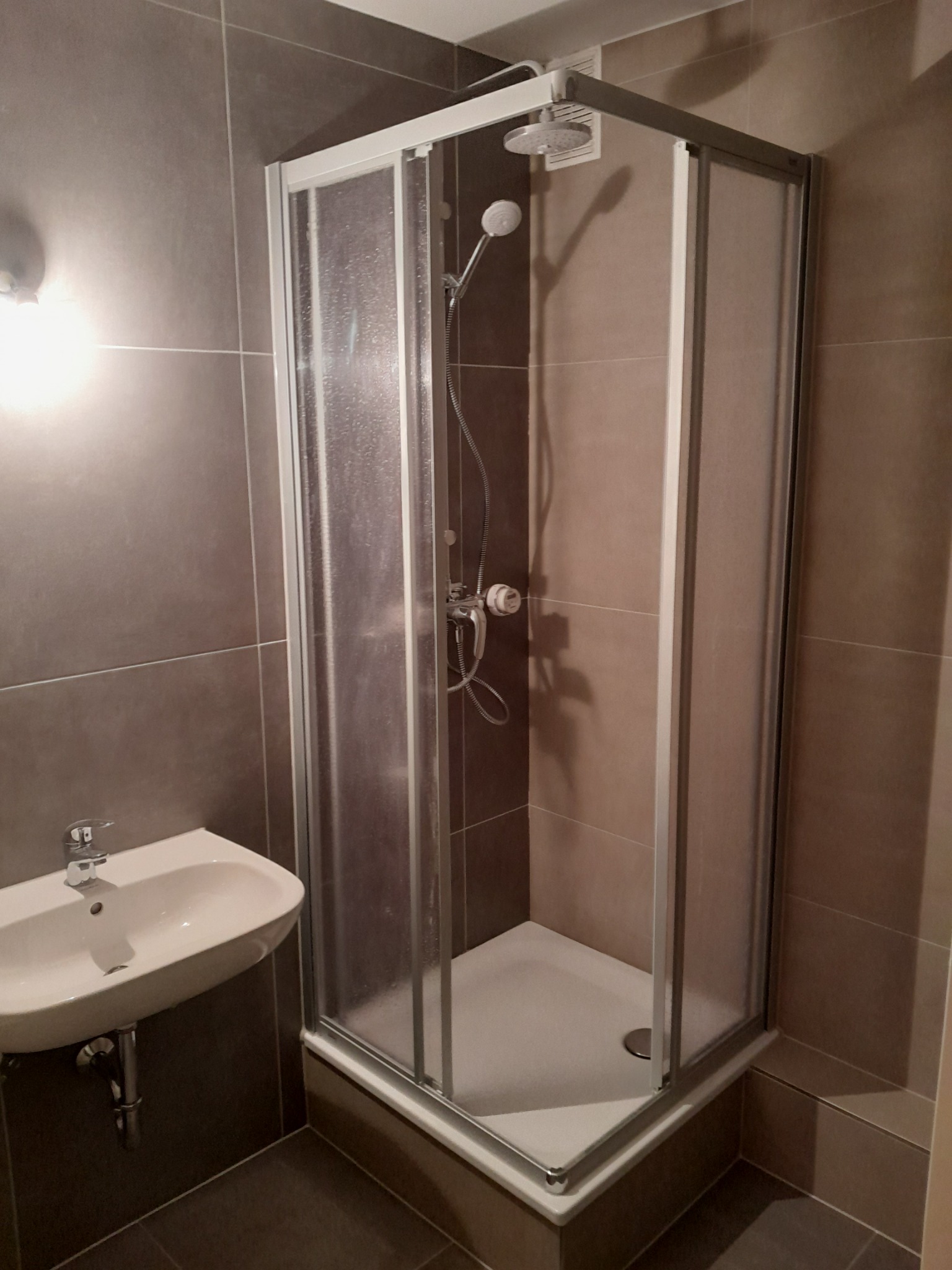 Narożna kabina prysznicowa z przesuwnymi drzwiami w łazience wyłożonej szarymi płytkami, umywalka ceramiczna zamontowana na ścianie z widocznym odpływem.