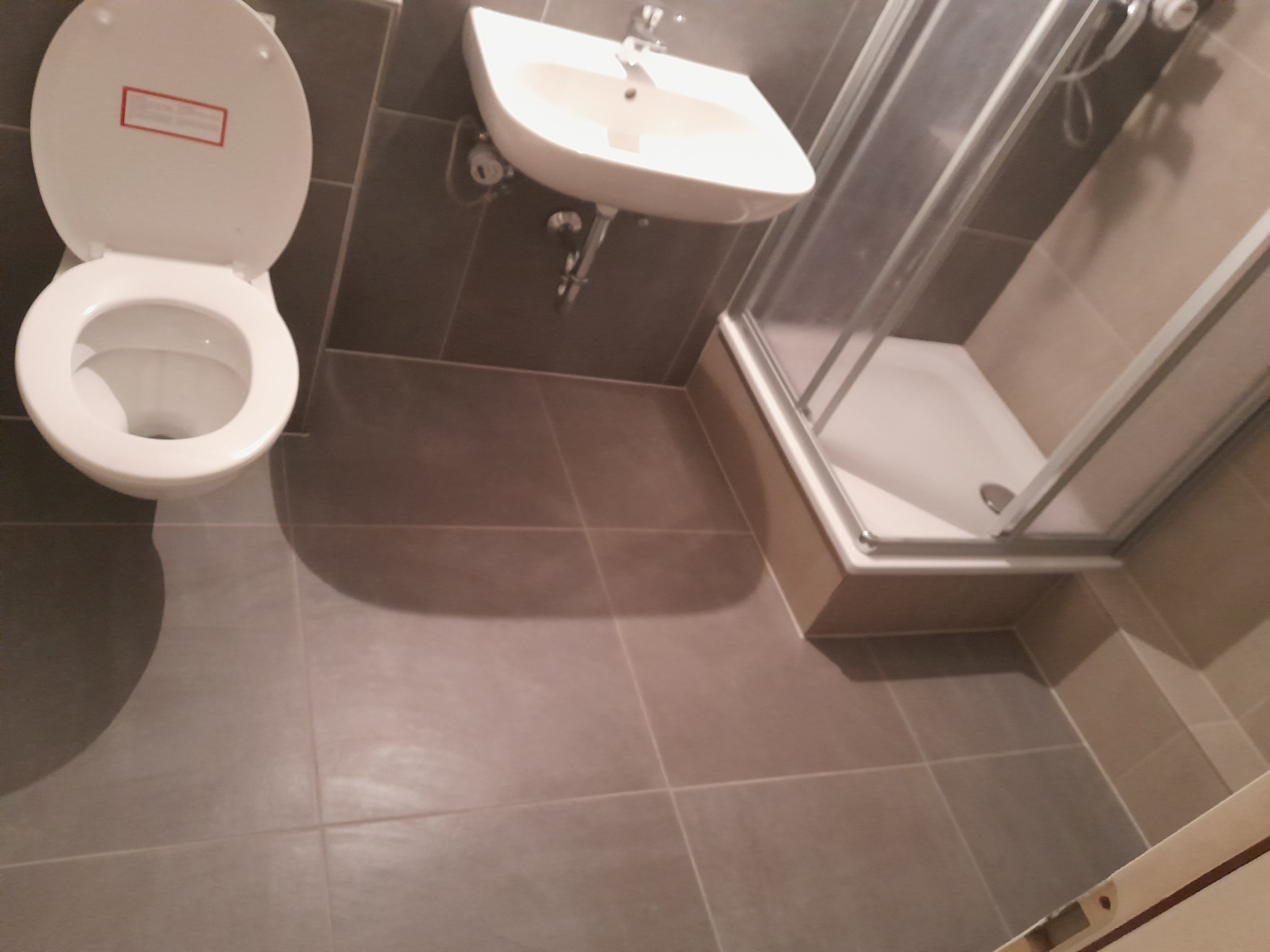 Widok z góry na wyremontowaną łazienkę z szarą glazurą, widoczny biały sedes z otwartą klapą, umywalka i kabina prysznicowa z aluminiowymi ramami.