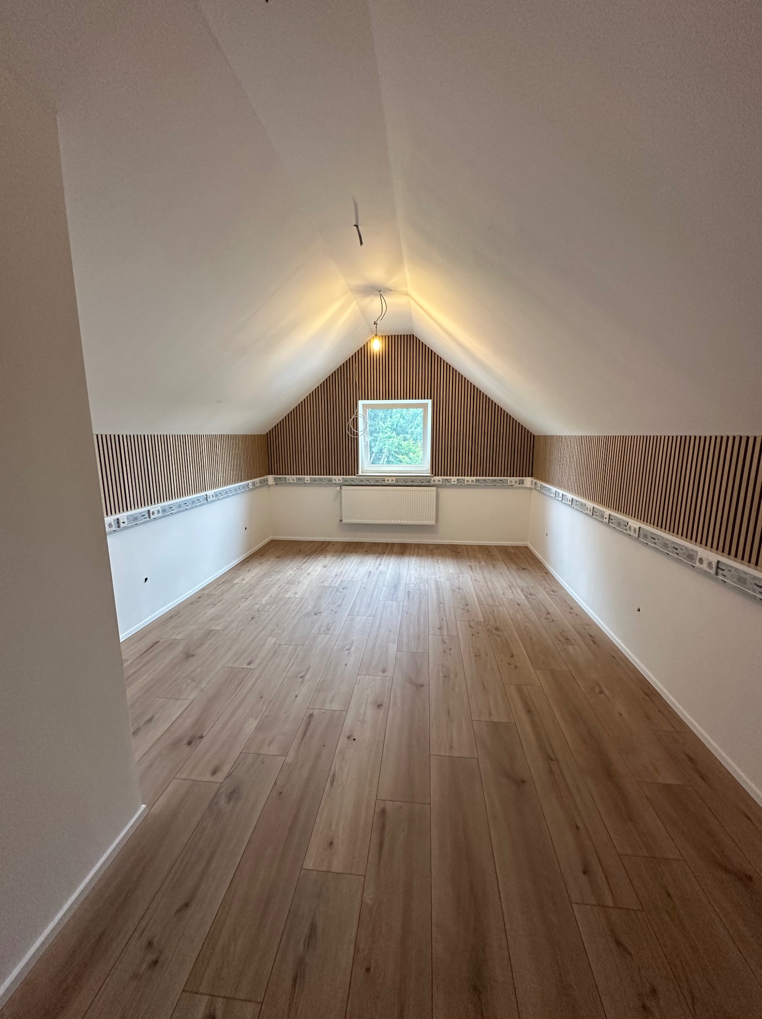 Poddasze z drewnianą podłogą i panelami ściennymi z pionowych listewek, widoczne szyny instalacyjne i okno dachowe.