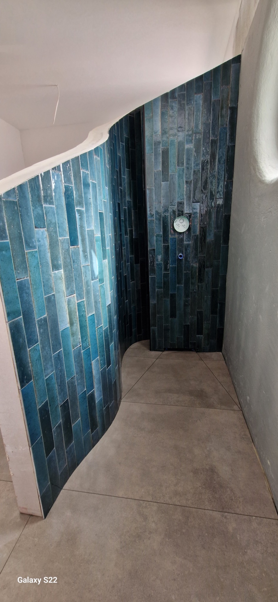 Niebiesko-turkusowa mozaika z płytek w kształcie cegiełek na zaokrąglonej ścianie prysznica z widoczną baterią podtynkową i szarą podłogą z płytek.