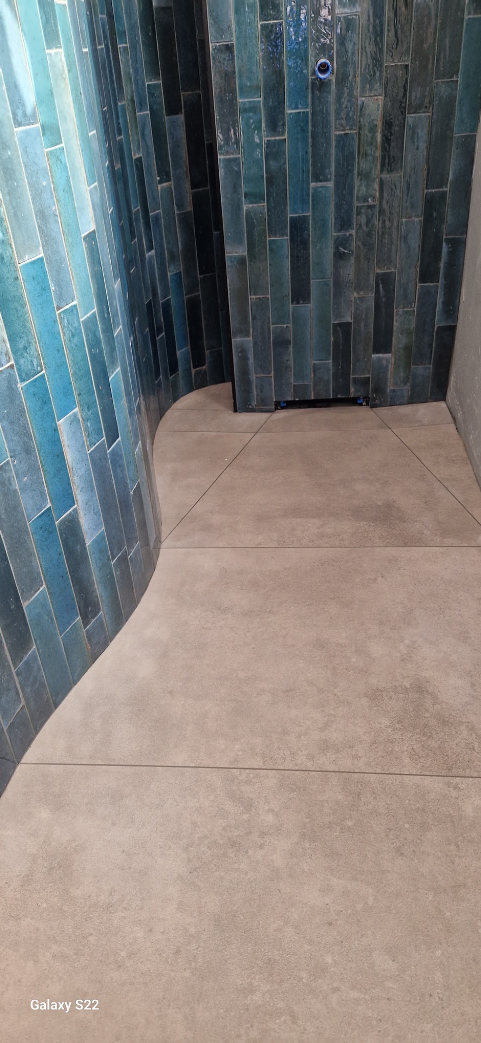 Ściana wyłożona pionowymi, turkusowymi płytkami o błyszczącej powierzchni, przechodząca w beżową, matową podłogę z płytek w łazience.