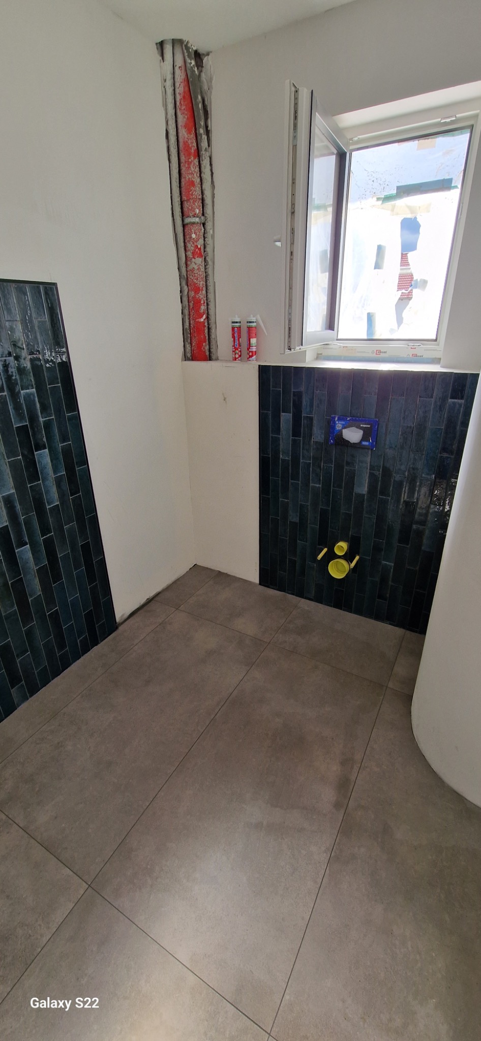 Łazienka w trakcie prac wykończeniowych: ułożone ciemne, pionowe płytki na ścianie, szare płytki podłogowe, przygotowane instalacje wodno-kanalizacyjne, widoczne rury w ścianie i okno.