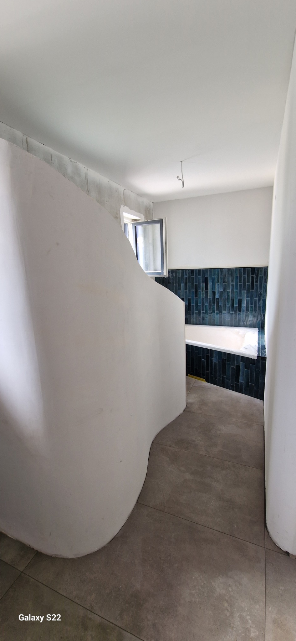 Łazienka w trakcie prac wykończeniowych: nietypowa, zaokrąglona ściana z surowym wykończeniem, ciemnozielona glazura na ścianie w tle, widoczna wanna i okno.