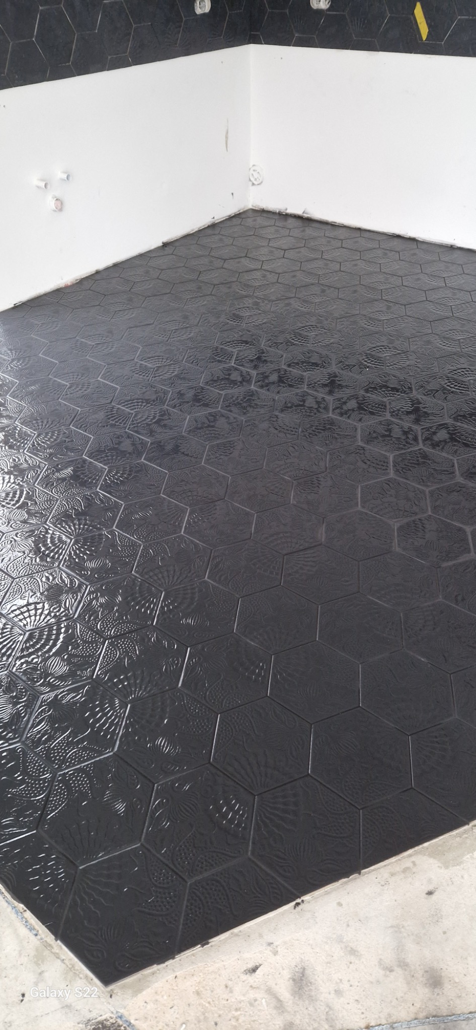 Ułożona, błyszcząca, czarna podłoga z heksagonalnych płytek z tłoczonym wzorem w narożniku łazienki z białymi ścianami i widocznymi instalacjami.