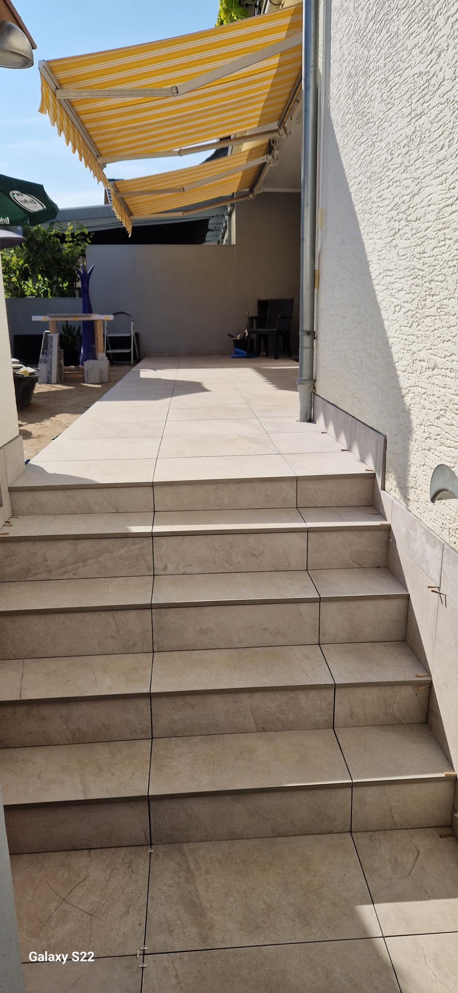 Wyłożone beżowymi płytkami schody prowadzące na taras z markizą w paski, widoczne plastikowe krzyżyki dystansowe w fugach.