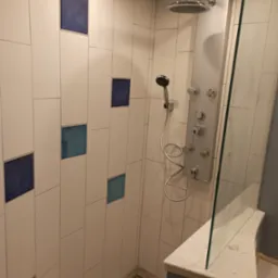 Nowoczesna kabina prysznicowa z białymi, prostokątnymi płytkami ułożonymi pionowo, z akcentami w postaci niebieskich kwadrat&oacute;w, panel prysznicowy z hydromasażem i deszczownicą, szklana ściana...