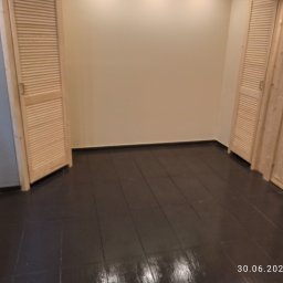 Rafał kozinski - Pomalowana na ciemny brąz podłoga z drewnianych desek w pokoju z beżowymi ścianami i dwojgiem jasnych drewnianych drzwi z żaluzjami, jedne z szybą.