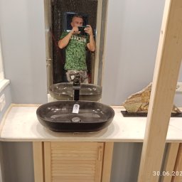 Rafał kozinski - Czarna, owalna umywalka nablatowa z chromowaną baterią, zamontowana na szafce z jasnego drewna z żaluzjowymi drzwiczkami, odbicie fotografa w lustrze.