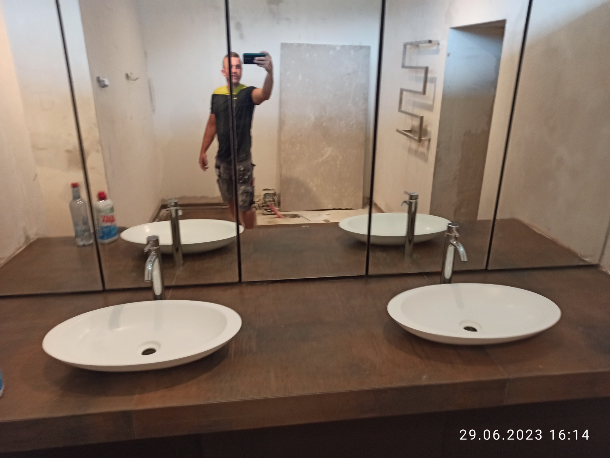 Łazienka w trakcie renowacji, widoczne dwie białe umywalki nablatowe z chromowanymi bateriami na blacie imitującym drewno, duże lustro odbijające osobę robiącą zdjęcie telefonem, ściany w trakcie...