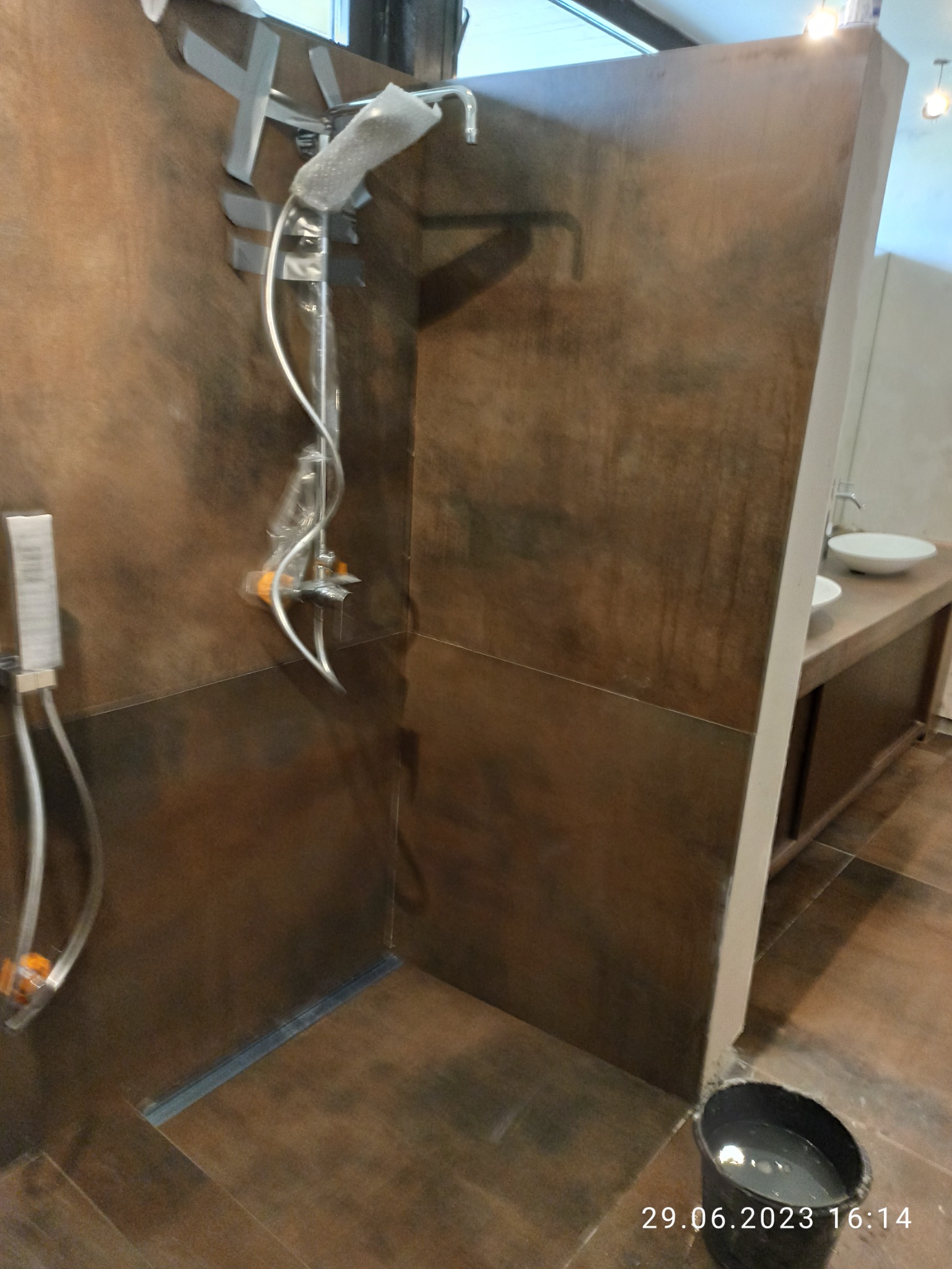 Niedokończony montaż baterii prysznicowej z folią ochronną i taśmą na brązowej ścianie w kabinie prysznicowej z odpływem liniowym, widoczne umywalki i wiadro z wodą.