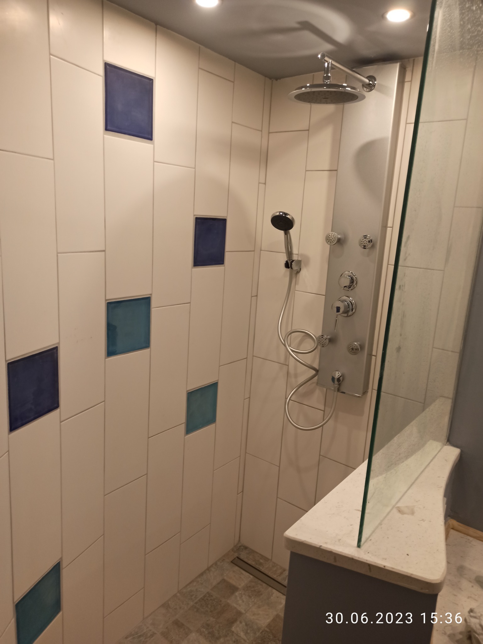 Nowoczesna kabina prysznicowa z białymi, prostokątnymi płytkami ułożonymi pionowo, z akcentami w postaci niebieskich kwadratów, panel prysznicowy z hydromasażem i deszczownicą, szklana ściana...