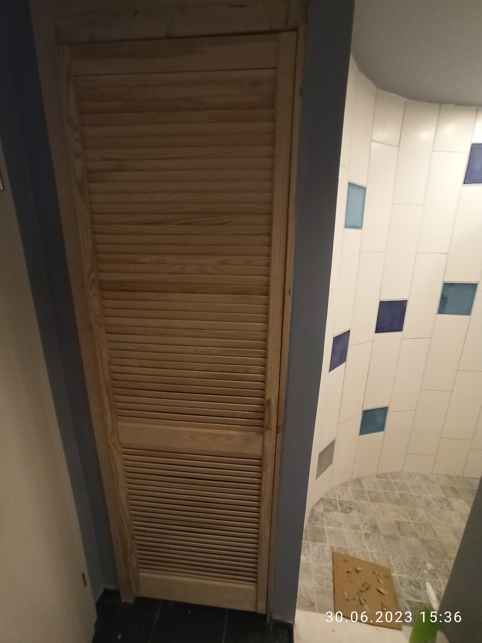 Zamknięte, drewniane drzwi lamelowe w łazience z białymi płytkami z niebieskimi akcentami. Widoczny fragment podłogi i ściany. Data i godzina na dole zdjęcia.