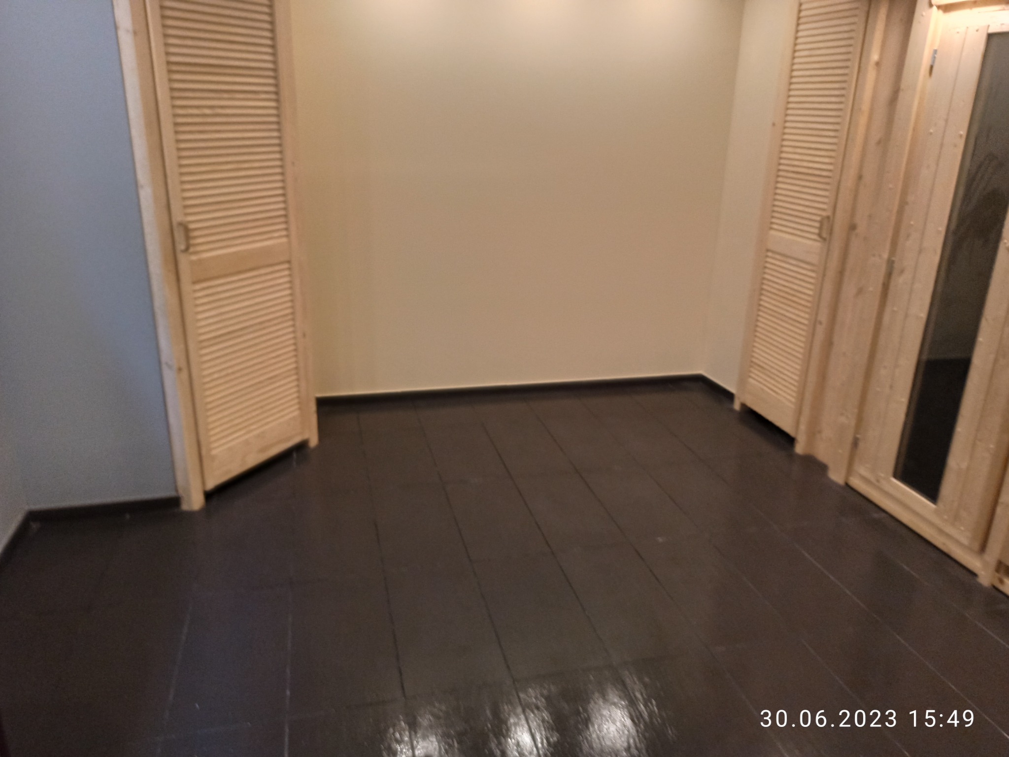 Pomalowana na ciemny brąz podłoga z drewnianych desek w pokoju z beżowymi ścianami i dwojgiem jasnych drewnianych drzwi z żaluzjami, jedne z szybą.