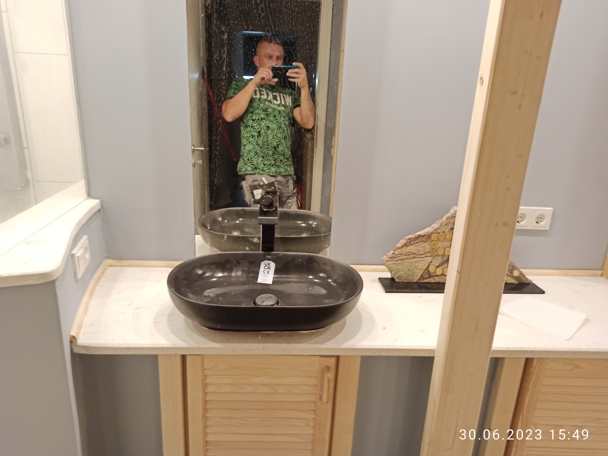 Czarna, owalna umywalka nablatowa z chromowaną baterią, zamontowana na szafce z jasnego drewna z żaluzjowymi drzwiczkami, odbicie fotografa w lustrze.