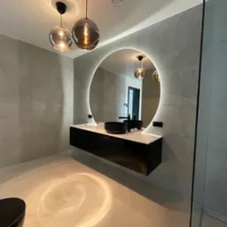 Nowoczesna łazienka z okrągłym lustrem podświetlanym LED, wisząca czarna szafka z białą umywalką, szare ściany z efektem betonu i szklana kabina prysznicowa.