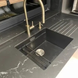 Czarny zlewozmywak wpuszczony w blat z ciemnego kamienia z białym żyłkowaniem, bateria i dozownik w kolorze szczotkowanego złota, ociekacz wkomponowany w blat.