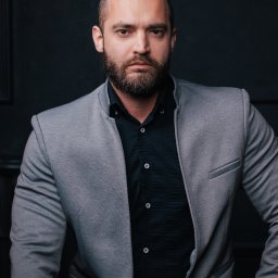 Paula Jankowska Photography - Portret mężczyzny z brodą w szarej marynarce i czarnej koszuli na tle ciemnej ściany, z tatuażem na dłoni.