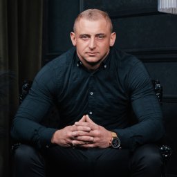 Paula Jankowska Photography - Portret mężczyzny w czarnej koszuli i spodniach, siedzącego na zdobionym krześle z rękami złożonymi na kolanach, z widocznym złotym zegarkiem na nadgarstku, na tle ciemnej ściany.