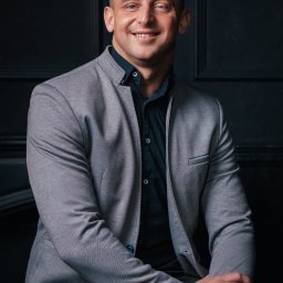 Paula Jankowska Photography - Uśmiechnięty mężczyzna w szarym garniturze i czarnej koszuli, z zegarkiem na bransolecie mesh i złotymi pierścieniami na palcach, pozuje na tle ciemnej, panelowej ściany.