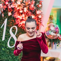 Paula Jankowska Photography - Uśmiechnięta kobieta w bordowej sukience na tle dekoracji z kwiatów i cyfry 18, unosząca palce w górę, widoczne złote kolczyki i pierścionki.