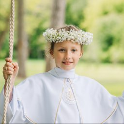 Paula Jankowska Photography - Dziewczynka w białej albie i wianku z białych kwiatów na huśtawce linowej, uśmiechnięta, z delikatnym makijażem, na tle zieleni drzew.