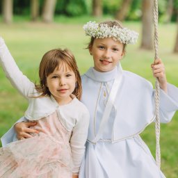Paula Jankowska Photography - Dwie dziewczynki na huśtawce w plenerze. Jedna w stroju komunijnym z wiankiem, druga w sukience w kwiaty. Uśmiechnięte, w naturalnym oświetleniu.