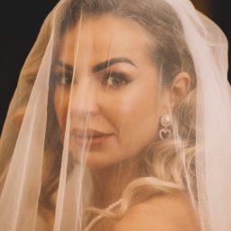 Paula Jankowska Photography - Portret panny młodej z delikatnym makijażem, ukrytej za woalką, z widoczną perłową biżuterią i kolczykami w kształcie serc na ciemnym tle.