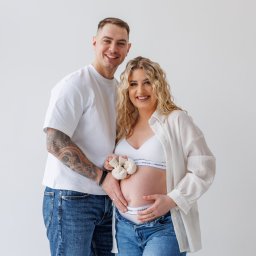 Paula Jankowska Photography - Uśmiechnięta para w jasnych ubraniach pozuje na sesji ciążowej. Kobieta trzyma małe, dziergane buciki na brzuchu. Mężczyzna ma tatuaż na ramieniu. Białe tło.