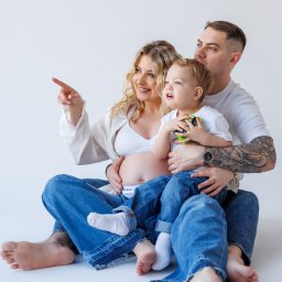 Paula Jankowska Photography - Uśmiechnięta, przyszła mama z blond lokami, tata z tatuażem i synek siedzą na białym tle. Kobieta wskazuje palcem w dal. Sesja w jeansach i bieli.